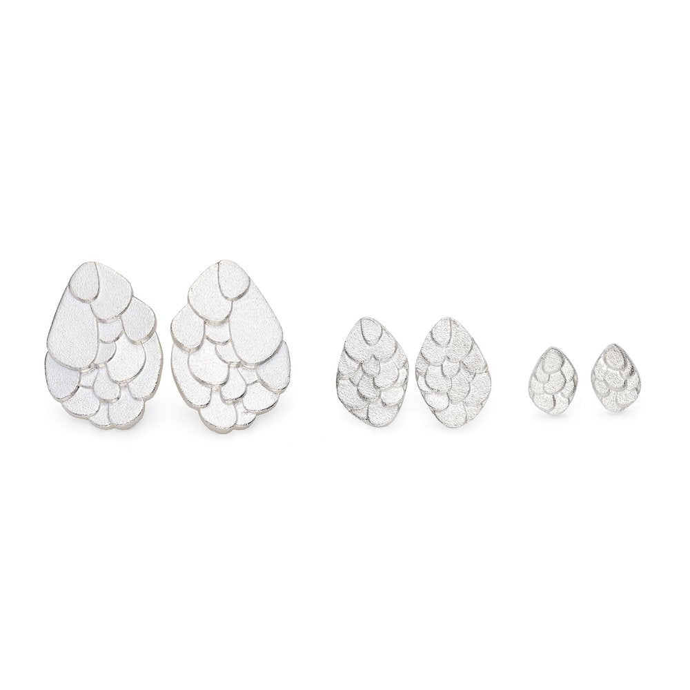 A set of Kimana Stud Earrings Silver by Leoma Drew featuring a teardrop shape with a layered, scalloped pattern. The set includes three pairs of frosted silver earrings in descending sizes, with the largest on the left and the smallest on the right. The design has a textured, shiny surface.