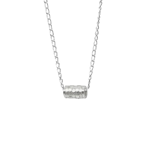 Here is the rewritten sentence with the product name and brand name included:
The Tiny Karma Wheel Necklace Silver by Andrea Mears Jewellery is a sterling silver necklace with a cylindrical, textured pendant hanging from a delicate chain. The cylinder, reminiscent of spinning prayer wheels, has a detailed, hammered surface that reflects light, adding a subtle shine to the design. The chain features evenly spaced links for an elegant look.