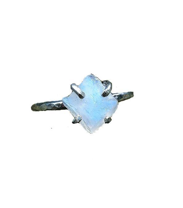 A close-up of the Caroline Stokesberry-lee Raw Moonstone Rock Ring featuring a raw, square-shaped moonstone set in a rustic, textured metal band with four prongs. The natural gemstone's pale blue and white hues reflect light, giving it an iridescent quality. The background is white and minimalistic.