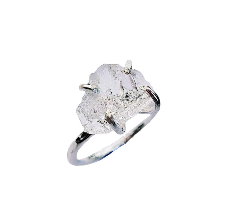 A sterling silver ring with a large, rough, uncut gemstone held by prongs is displayed against a white background. The Raw Quartz Rock Ring by Caroline Stokesberry-lee has a clear, crystalline appearance, and its raw, natural texture contrasts beautifully with the smooth finish of the silver band.