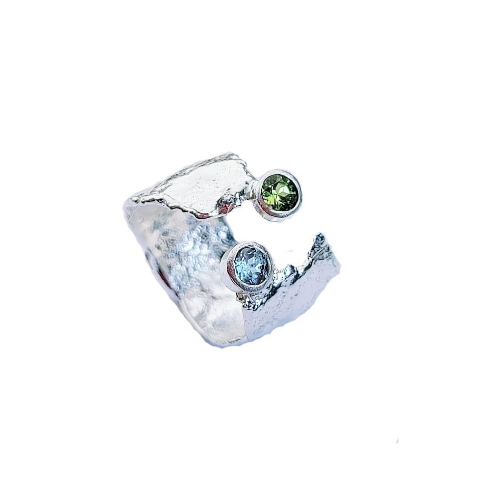 A rustic sterling silver Caroline Stokesberry-lee Blue Topaz & Peridot Faerie Tale Ring features a unique, textured band with Blue Topaz and Peridot stones. The open-ended design, with one green stone and one blue stone set at the ends, adds a touch of color and elegance to the rugged design. The background is plain white.