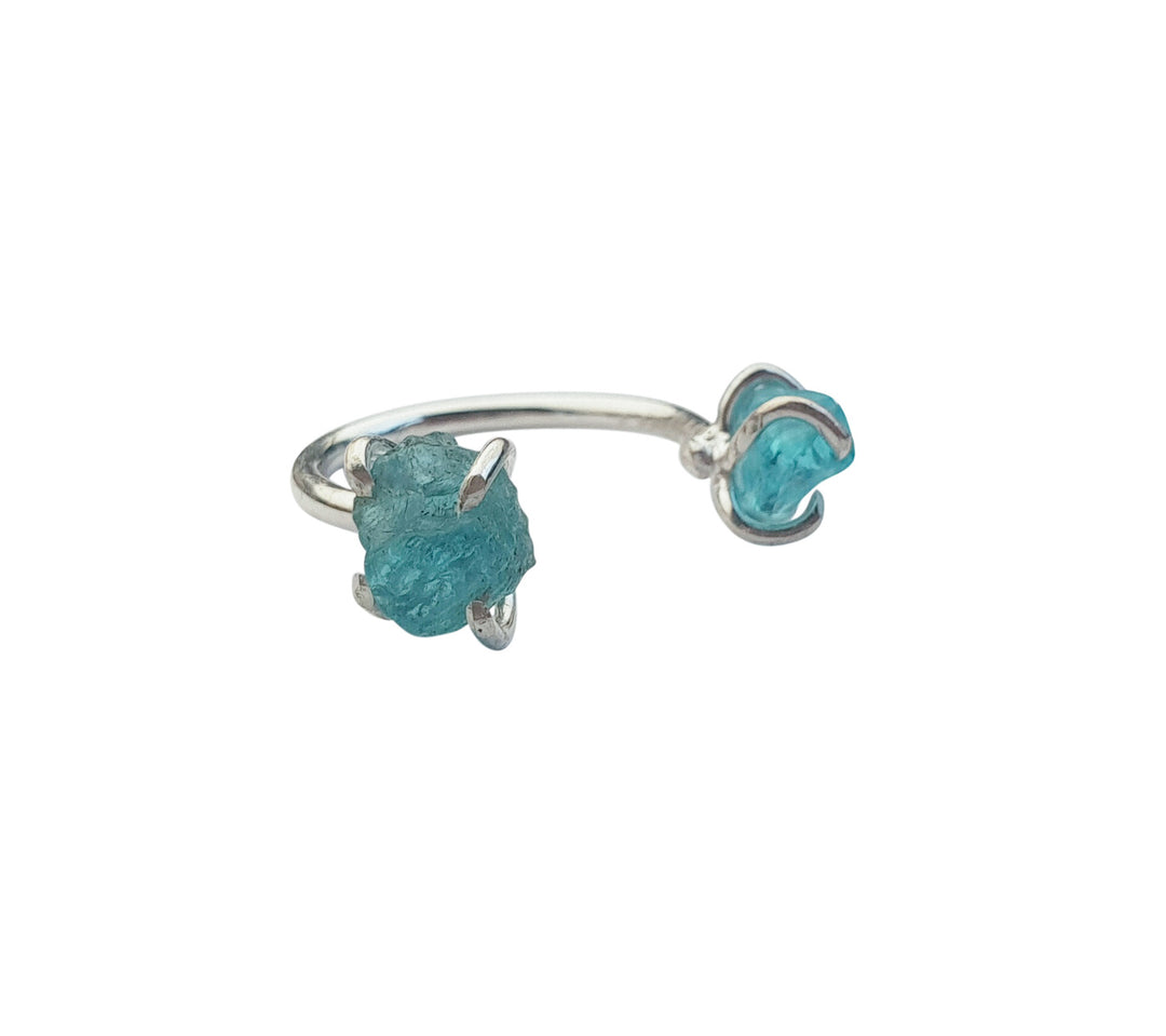 An open-ended silver ring featuring two blue raw stone crystals, each set in pronged settings at either end. One stone is slightly larger and more irregularly shaped than the other, giving the ring a modern, asymmetrical look. This is the Twin Flame Ring Sterling Silver & Raw Blue Apatite by Caroline Stokesberry-lee.