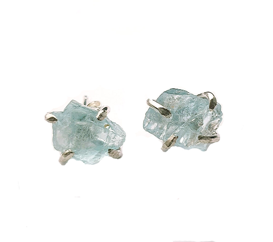 Two Raw Aquamarine Stud Earrings by Caroline Stokesberry-lee featuring raw Aquamarine gems set in sterling silver claws. The earrings have an organic, asymmetrical appearance, showcasing the natural shape and texture of the stones. Sterling silver ear posts complete the design, with a white background highlighting the earrings.