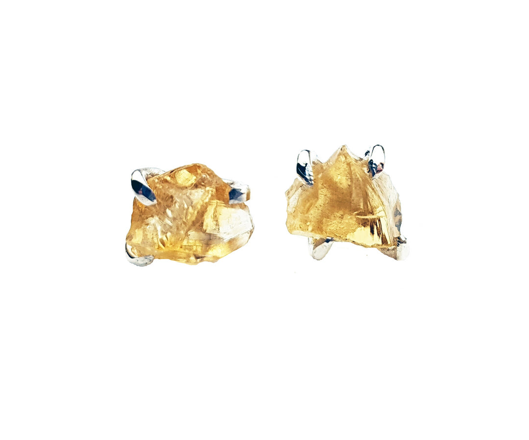 A pair of Caroline Stokesberry-lee Raw Citrine Stud Earrings featuring uncut, raw citrine gemstones set in minimalist sterling silver prongs. Each stone has a rough and irregular shape, highlighting its natural texture and color. The earrings are placed on a white background.