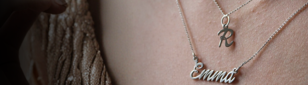 Two silver name necklaces, one reading "Sarah" and the other "Emma," hang on chains against a plain gray background with the words "Name Necklaces" to the right.