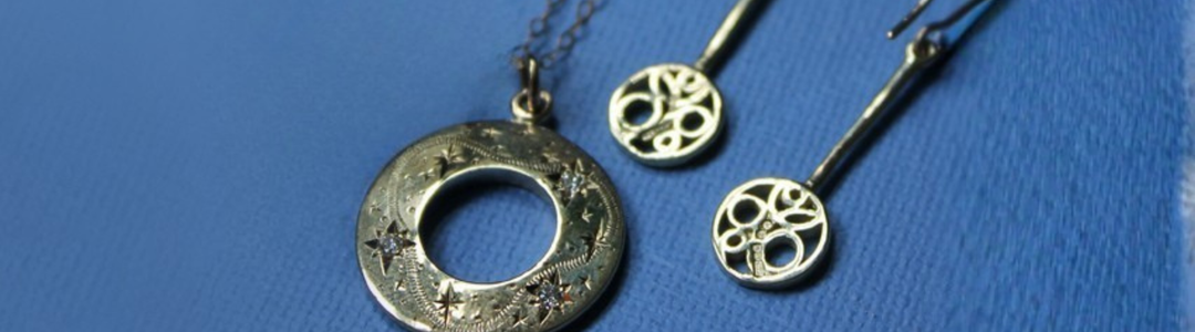 A close-up of elegant silver jewelry on a blue surface, including a ring with a circular design, a pendant necklace, and two matching earrings with intricate detailing.