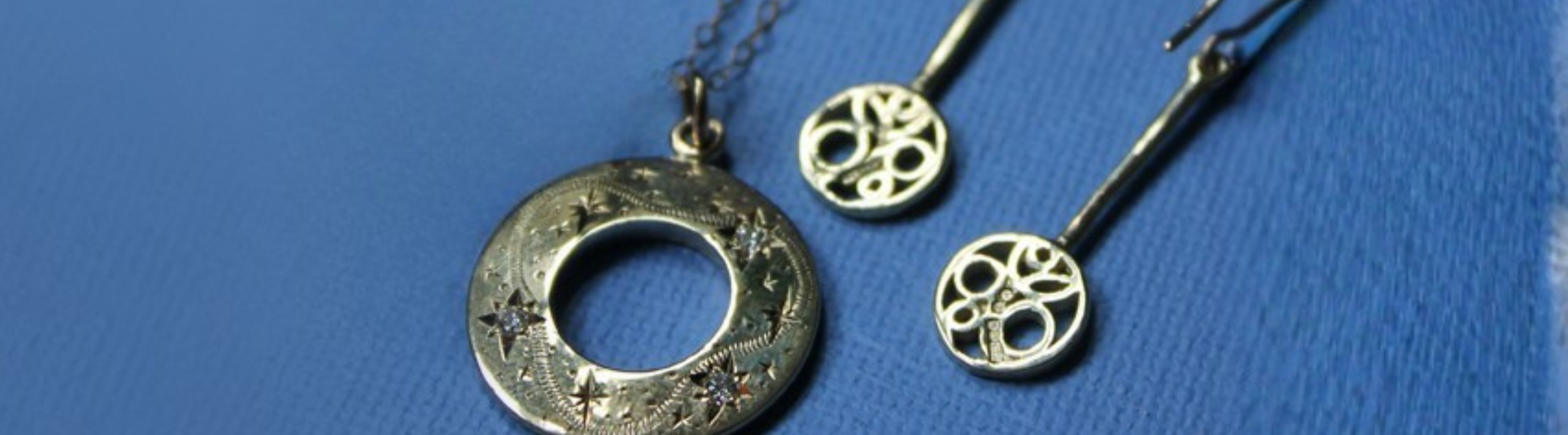 A close-up of elegant silver jewelry on a blue surface, including a ring with a circular design, a pendant necklace, and two matching earrings with intricate detailing.