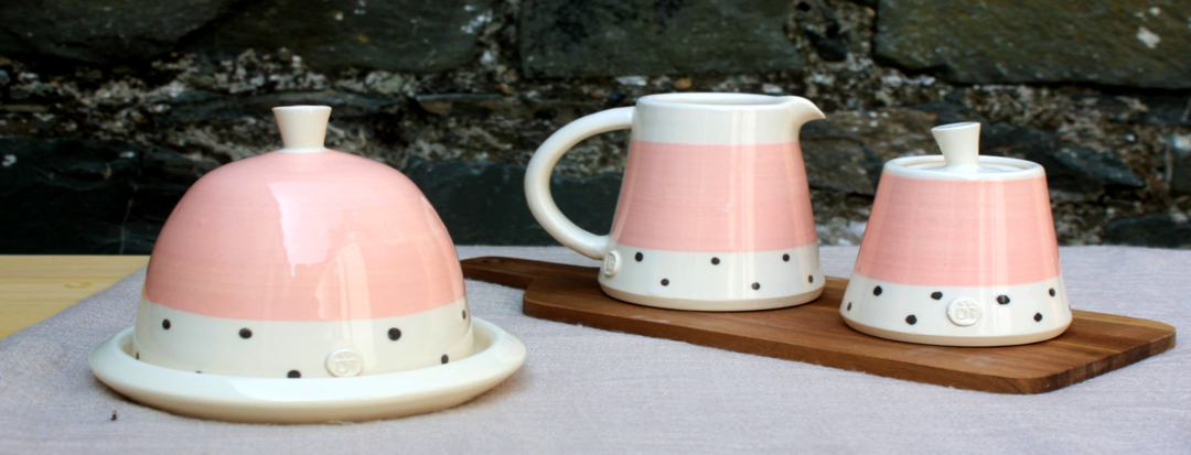 A set of ceramic kitchenware featuring a pink and white polka dot pattern, including a butter dish, a cream jug, and a sugar bowl, all placed on a wooden tray against a stone background.