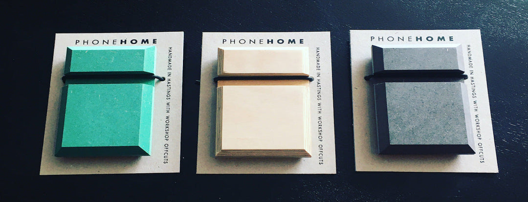 Three rectangular phone stands in teal, light wood, and dark wood are displayed side by side on cards labeled PHONEHOME. The cards also mention that the stands are handmade in Hastings with workshop offcuts.