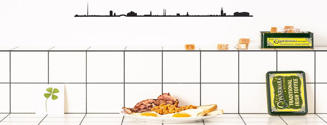 A plate of traditional Irish breakfast including eggs, beans, and meat sits on a white tiled counter. Beside it, there's a tin of Connemara Irish Toffee and a shamrock card. A skyline silhouette decorates the wall.