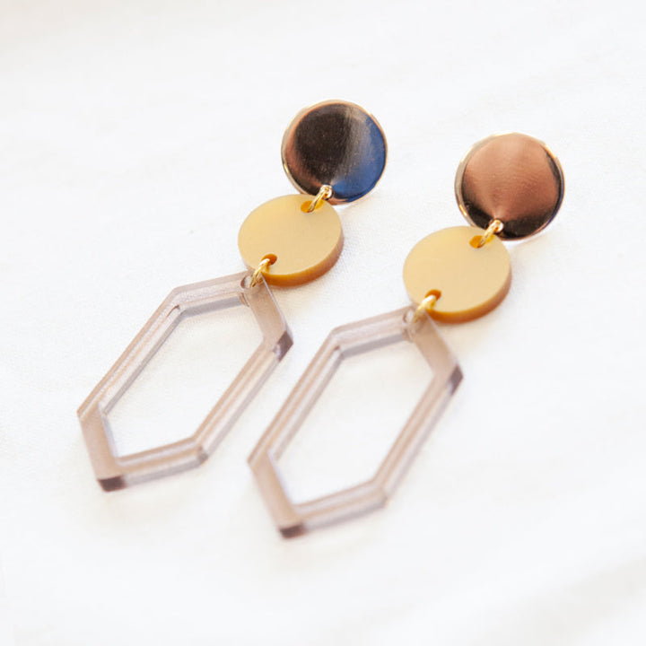 A pair of handmade Soul 04 Earrings by Studio Nok Nok on a white surface. Each earring features a round gold-plated top, a yellow circular middle piece, and a geometric, recycled acrylic hexagonal drop at the bottom. The design epitomizes modern and minimalist style.