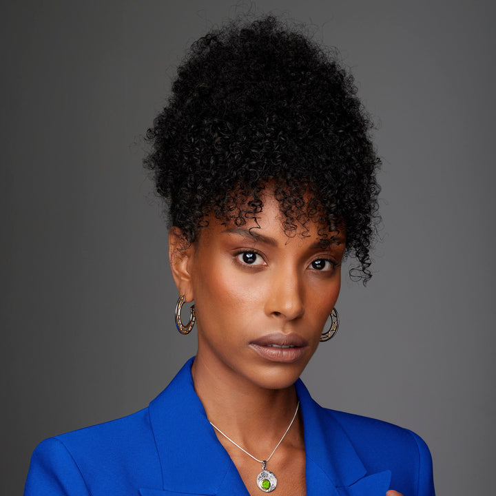 A woman with curly hair styled in an updo is wearing a royal blue blazer, **Maya Earrings by Gallardo & Blaine Designs**, and a necklace with a sterling silver pendant. She has a neutral expression and is set against a plain gray background.