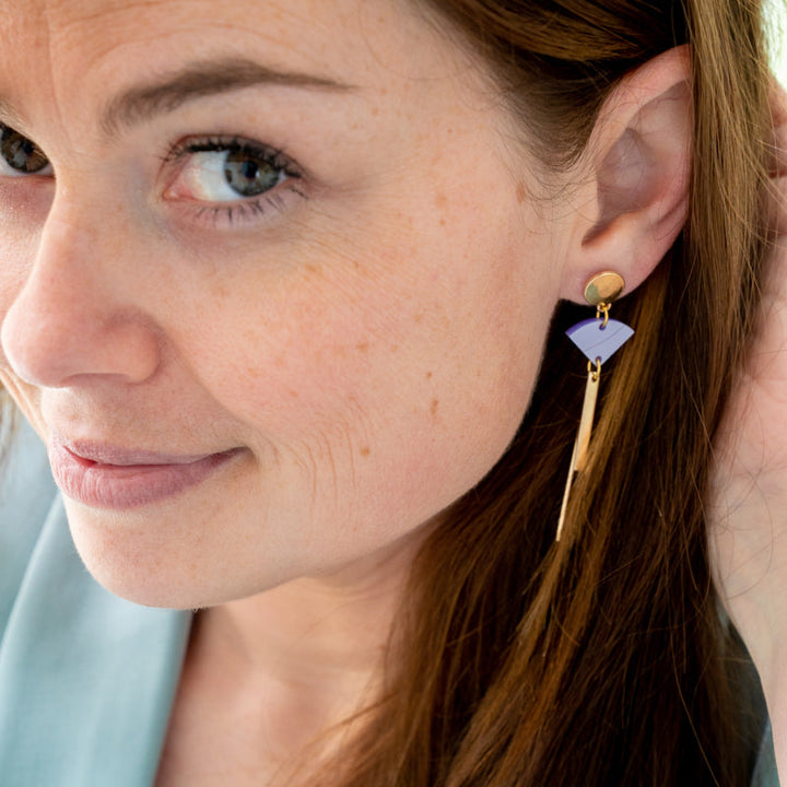 Close-up of a person with long brown hair smiling slightly. They are wearing a dangling gold-plated earring with a purple, triangular detail from Studio Nok Nok's "Sun 08 Earrings". The person is wearing a light-colored top, and their hand is gently touching their hair near the ear.