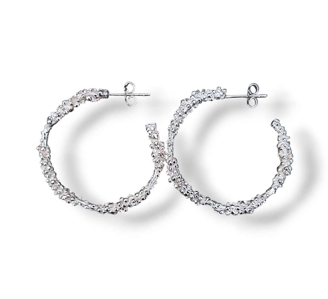 The Starborne Sterling Silver Hoop Earrings by Pale Raven feature a textured finish and post backs. Handcrafted in Dublin, these hoops are displayed on a white background with soft shadows underneath. No stones included.