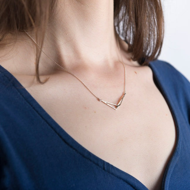 A close-up of a woman wearing the Miriam Wade Le Cheile Pendant Small Silver, a dainty sterling silver pendant in a geometric, V-shaped design. She is dressed in a deep blue, V-neck top, and her brown hair is partially visible, falling to her shoulders. The background is blurred, focusing on the necklace.