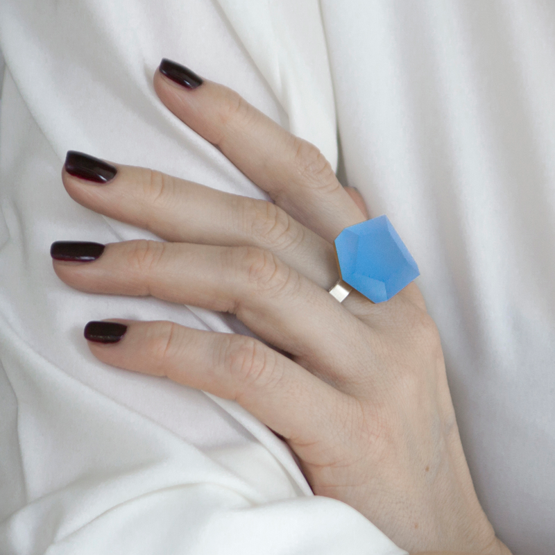 A hand with dark red nail polish is resting on a white fabric. The person is wearing a sterling silver ring with a large, geometric blue gemstone on the middle finger— the Fruit Bijoux Ring Top VU Crystals Sky Blue from Fruit Bijoux.