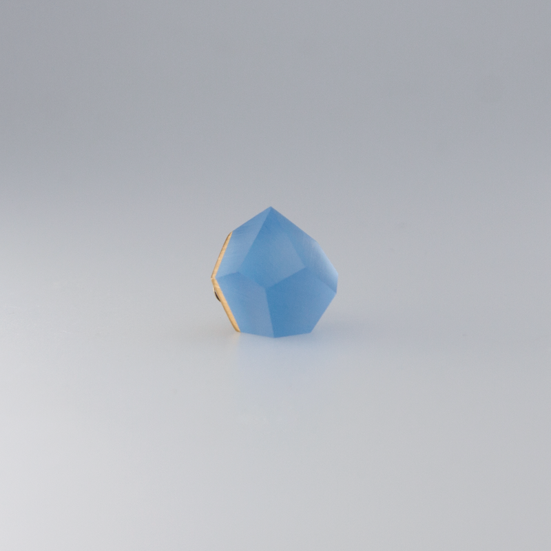 A small, geometric-shaped blue gemstone with multiple facets is displayed against a simple white background. The Fruit Bijoux Fruit Bijoux Ring Top VU Crystals Sky Blue, set as a ring top on sterling silver, has a slightly glossy surface, and its edges and angles are clearly defined.