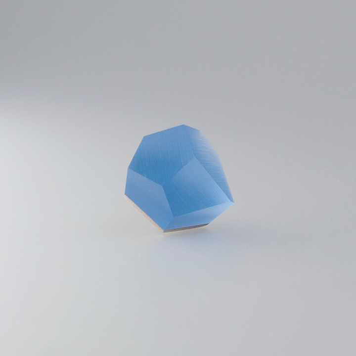 A single, faceted, Fruit Bijoux Ring Top VU Crystals Sky Blue gemstone rests on a plain white surface. It has multiple sharp edges and a glossy finish, reflecting light from its facets. The background is gradient, transitioning from light gray at the bottom to white at the top—a perfect centerpiece for any sterling silver ring top.