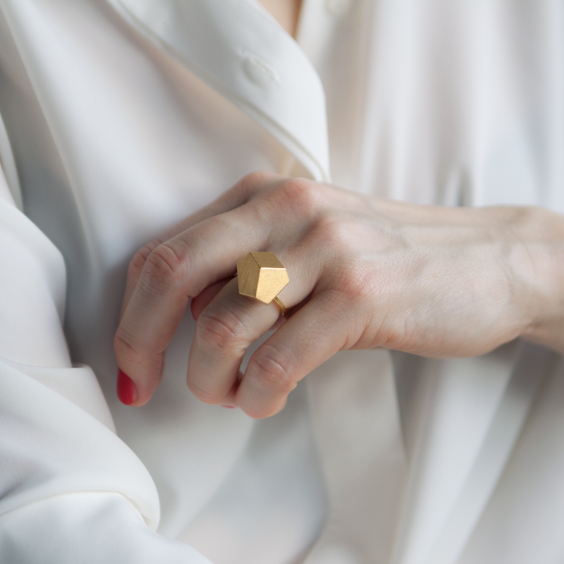 Close-up of a person wearing a white shirt, showcasing their hand adorned with a Fruit Bijoux Ring Top Goldrush by Fruit Bijoux and red nail polish on their pinky finger. The background is soft and blurred, emphasizing the handcrafted beauty of the piece.