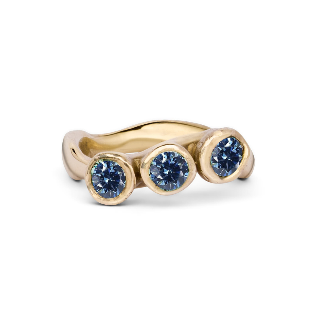 The Moonwater Ring by Madeleine Sculptural Jewellery is a handmade gold ring with three round blue stones in bezel settings and a gently wavy band—an elegant, hand-carved piece.