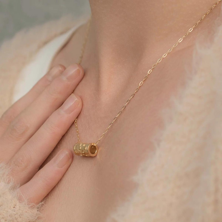 A close-up of a person wearing an Andrea Mears Jewellery Karma Wheel Necklace Gold with a cylindrical gold pendant. The person's hand is gently touching their chest near the pendant. The person is wearing a light-colored, fuzzy garment, highlighting the delicate allure of the gold filled chain.