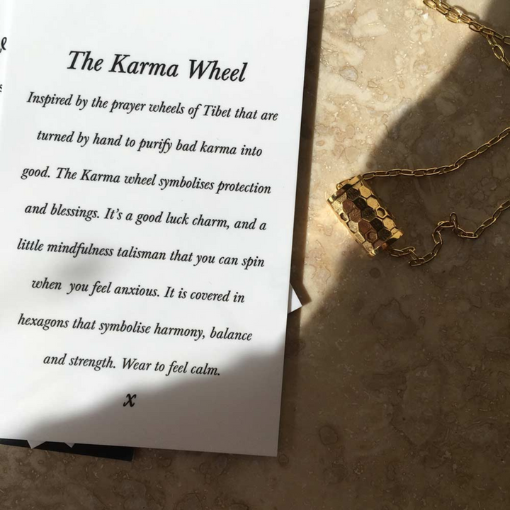 A pamphlet titled "The Karma Wheel" rests on a beige textured surface. The text explains the symbolism and purpose of the **Karma Wheel Necklace Gold from Andrea Mears Jewellery**. Beside it, a gold filled chain with a hexagon-patterned charm glimmers delicately. Shadows cast by the items are elegantly visible.