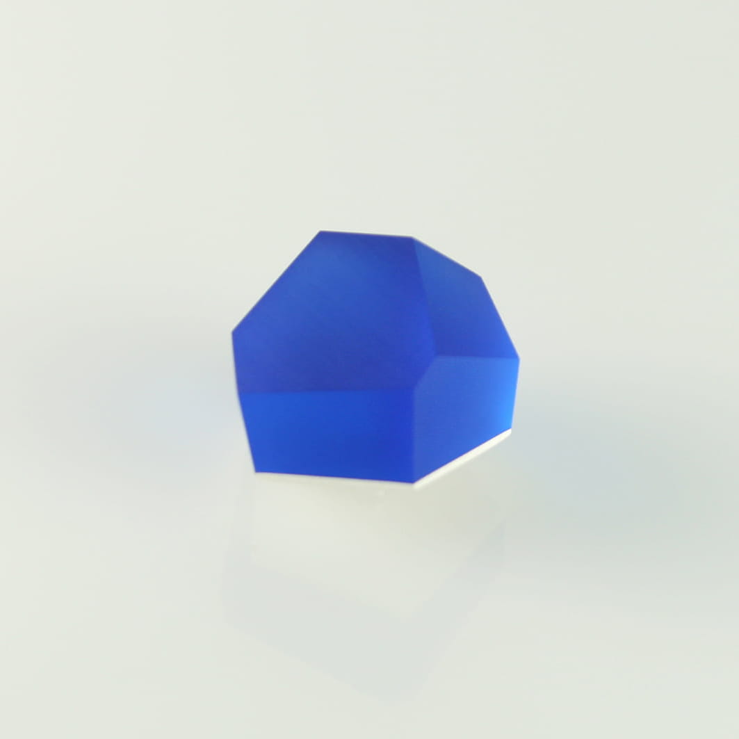 A Fruit Bijoux Ring Top VU Crystals Cobalt Blue with a matte finish is displayed against a white, softly-lit background. The Fruit Bijoux crystal, resembling a striking ring top, has an irregular, multifaceted shape and casts a faint reflection on the surface.