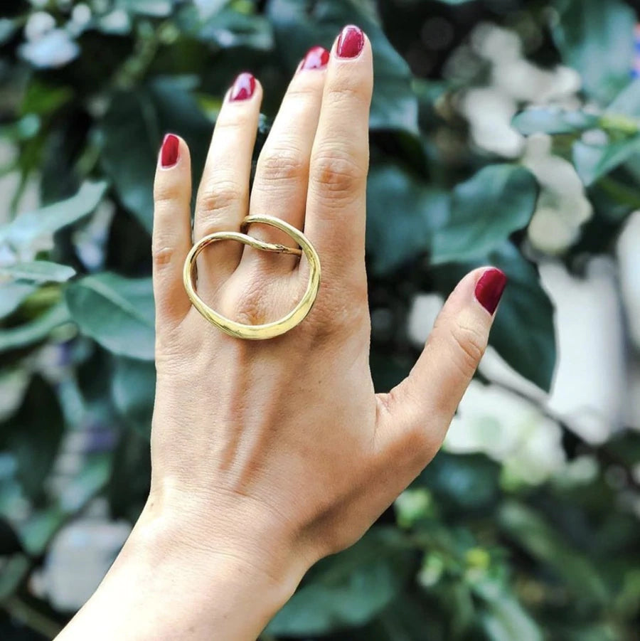 A hand with red nail polish is shown in front of a green leafy background. The middle finger is adorned with a large, gold-plated brass ring, the Infinity Ring Gold, fashioned by Lotta et Djossou.