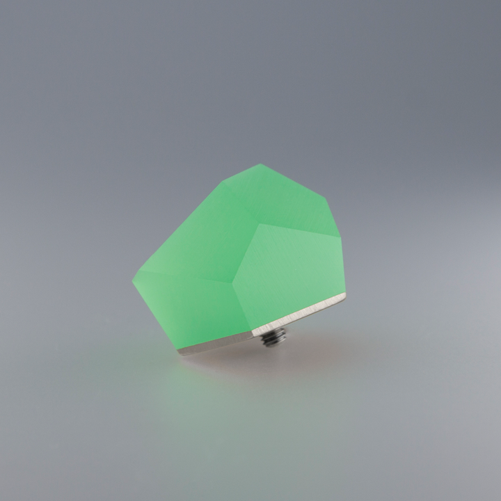 A minimalist image featuring a green, geometric, faceted shape resting on a smooth, light gray surface. The object has a metallic base with a small screw attachment, and its ring top is crafted from sterling silver, giving it a modern and sleek appearance. This is the Fruit Bijoux Ring Top VU Crystals Flash Green by Fruit Bijoux.