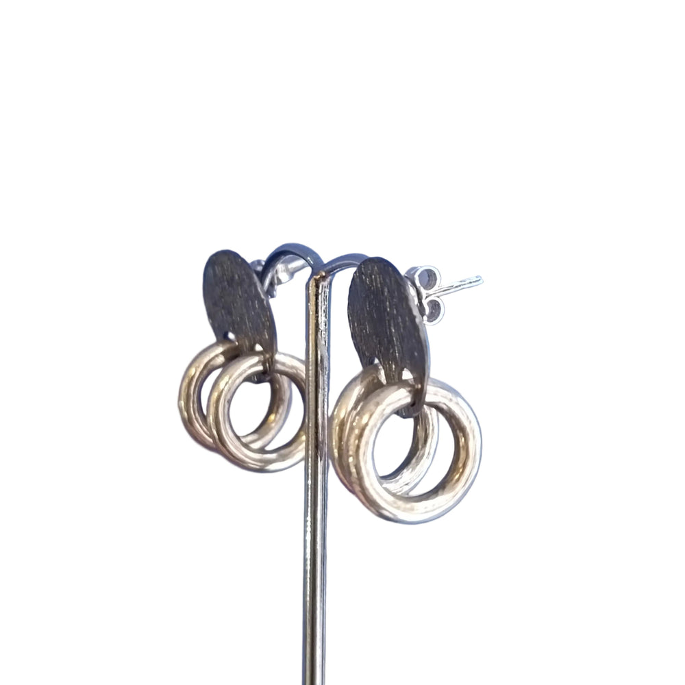 The K.And Double Ring Stud Earrings feature oval black studs and two interlocking gold rings, displayed on a metal stand against a blue background.
