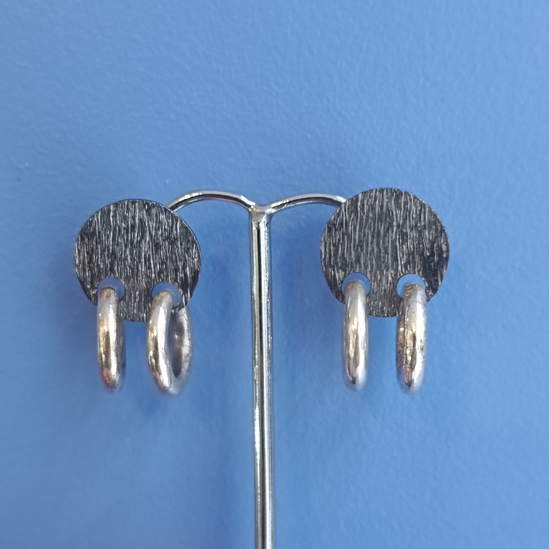 The K.And Double Ring Stud Earrings, featuring textured silver oval-shaped double hoops, are displayed on a metal stand against a blue background.