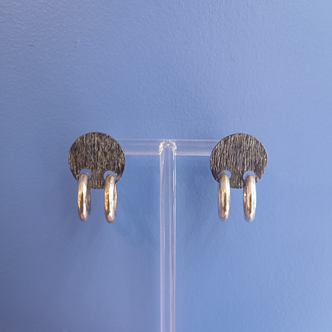 The K.And Double Ring Stud Earrings feature black oval bases with two small textured silver hoops on each, displayed on a clear stand against a blue background.