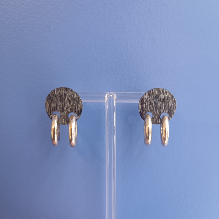 The K.And Double Ring Stud Earrings feature black oval bases with two small textured silver hoops on each, displayed on a clear stand against a blue background.