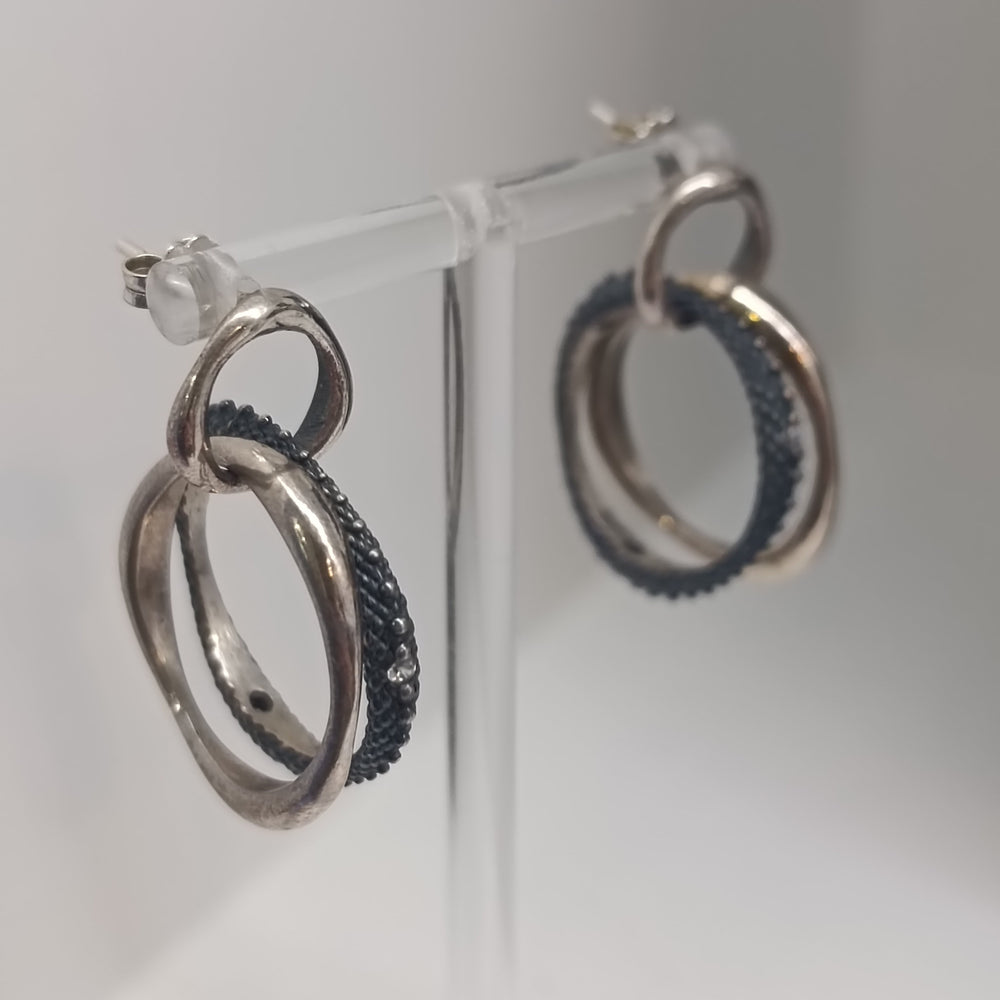 The K.And Textured & Oxidised Interlocking Circle Earrings with CZ feature two sterling silver rings—one smooth, one textured with black cubic zirconia—and are shown on a clear stand against a plain background.