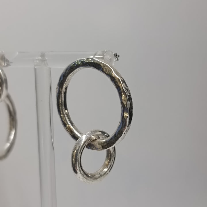 A close-up of K.And's Asymmetrical Chunky Silver Circle Earrings, showcasing two hammered, interlocking hoops of different sizes, displayed on a clear stand against a plain light gray background.