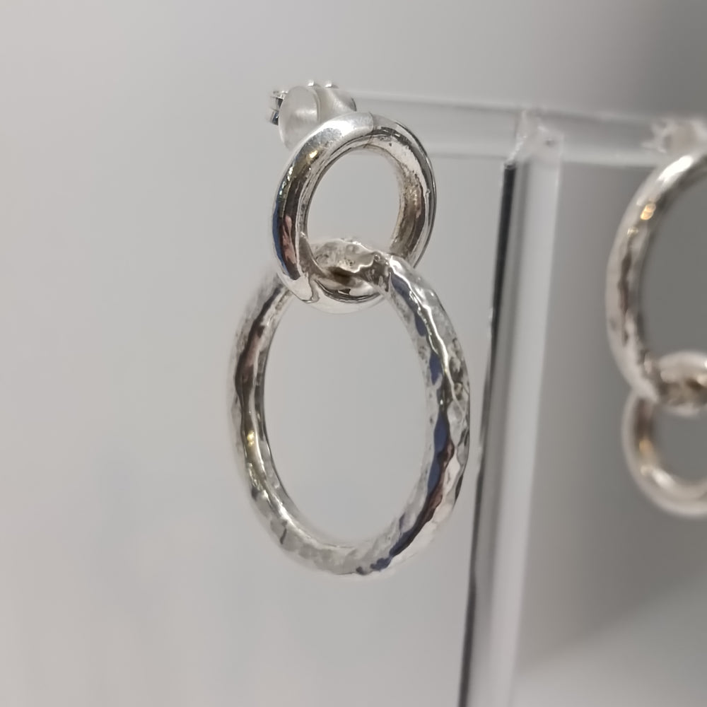 Close-up of K.And Asymmetrical Chunky Silver Circle Earrings featuring two interlocking rings—one small and smooth atop a larger textured one—displayed on a clear stand against a plain light background.