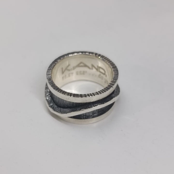 The Wave Spinner Ring by K.And features a textured silver design with a darkened oxidized center band and engraved “KAND” and “925” inside. This unique sterling silver ring is shown on a white surface.