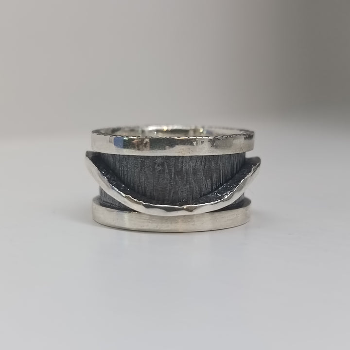 The K.And Wave Spinner Ring features a handcrafted textured sterling silver band with a wavy, polished edge and oxidised finish. This unique ring wraps partially around the center and is displayed on a plain white surface with a blurred background.