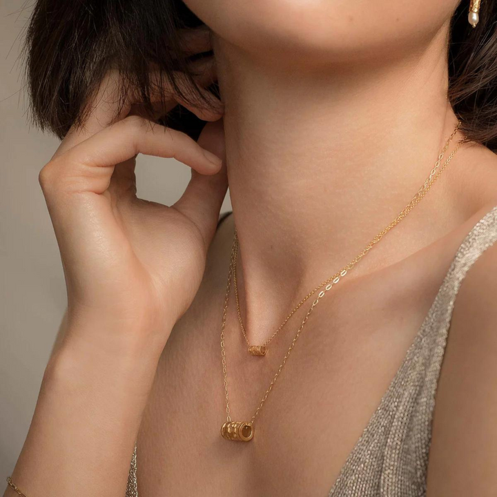 A person wearing two delicate gold necklaces, one of which is a Tiny Karma Wheel Necklace Gold by Andrea Mears Jewellery with an 18kt gold filled chain, is shown. The person is touching their face with their hand and is dressed in a sleeveless, shimmery top. The neutral background highlights the stunning jewelry.