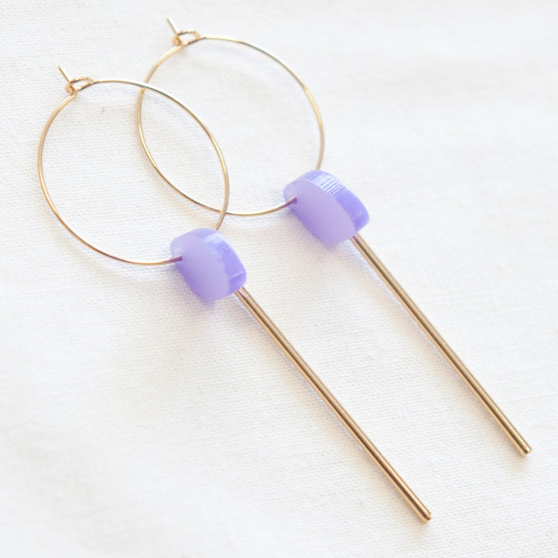 A pair of Studio Nok Nok Smile 06 Earrings with gold-toned hoops crafted from gold-plated nickel-free brass. Each hoop features a small, violet bead and a sleek, straight gold rod dangling from the bead. The earrings rest elegantly against a white fabric background.