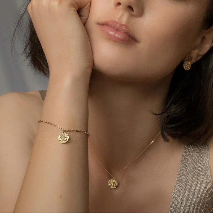 A woman wearing a sleeveless top showcases gold jewelry, including the Queen Bee Necklace Gold by Andrea Mears Jewellery, a bracelet, and earrings, each featuring a delicate coin pendant with a floral design reminiscent of an ancient Greek coin. The image focuses on her face, neck, and shoulder, emphasizing the elegant jewelry.