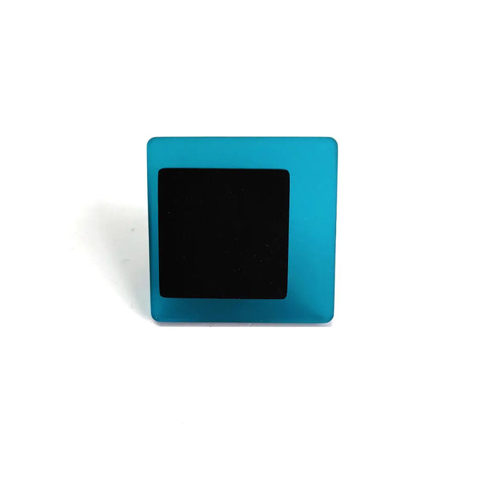 A small, square-shaped object with a teal blue frame and a black center is placed against a plain white background. Evoking the contemporary touch of the INFINITE LINE collection, the Eriko Ring by Alex + Svet is positioned upright and casts a faint shadow to the left.