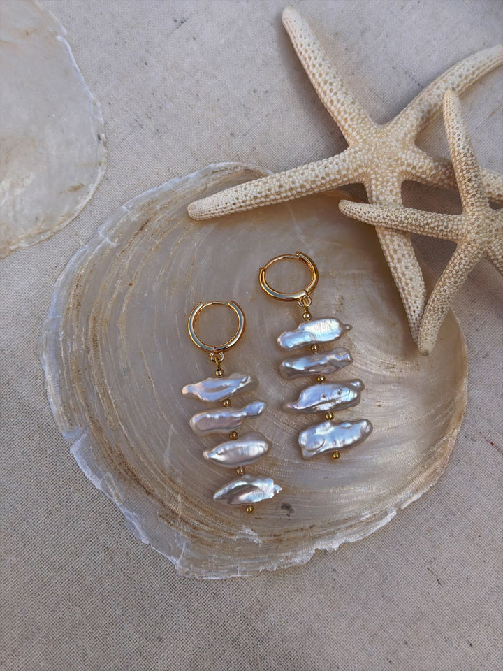 The Perla Sol Earrings by Kybalion Jewellery, featuring gold plating and stacked biwa pearls, are displayed on a translucent shell with starfish and a partial shell on a neutral fabric background.