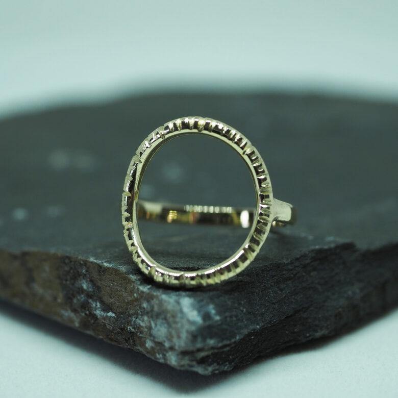 An Into The Light Ring 9ct Yellow Gold by Aud rests on a rough, dark-colored stone slab. The background is a soft, blurred gradient of light colors, drawing focus to the ring's intricate detailing and the contrasting surface it is placed on.