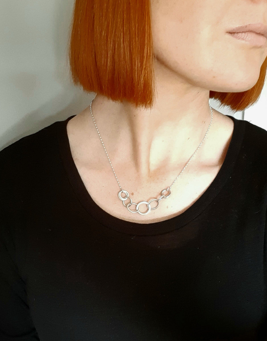 Close-up of a person with short, straight red hair, wearing a black top and a sterling silver Carran Chain Necklace by Lynsey de Burca with a design of interconnected circles. The person's face is partially visible, turned slightly to the right.