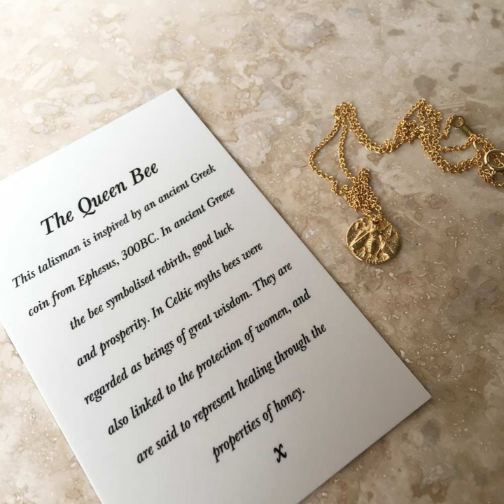 A Queen Bee Necklace Gold from Andrea Mears Jewellery, inspired by an ancient Greek coin, lies on a marble surface next to a card titled "The Queen Bee." The card explains the talisman’s meaning: a power necklace symbolizing rebirth, luck, prosperity, wisdom, protection of women, and honey’s healing properties.