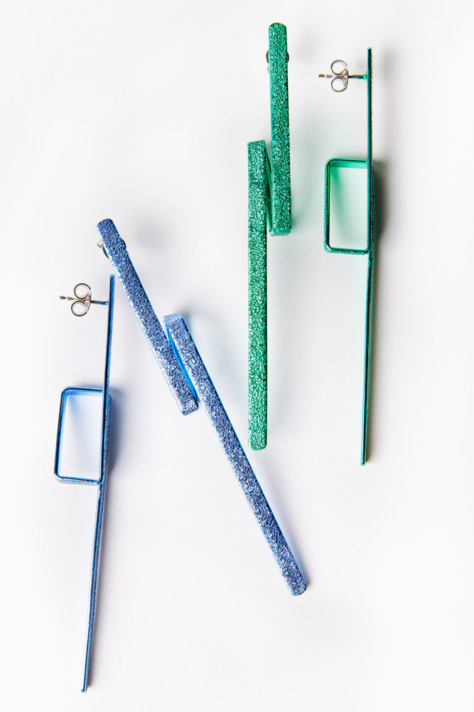 Two pairs of drop earrings are displayed against a white background. One pair, the Lysithea Green Earrings by Theodora D, is green with square and linear shapes, adorned with a green ceramic coating, while the other pair is a similar design but in blue. Both pairs have a minimalist and geometric style, crafted from 925 sterling silver for an elegant touch.