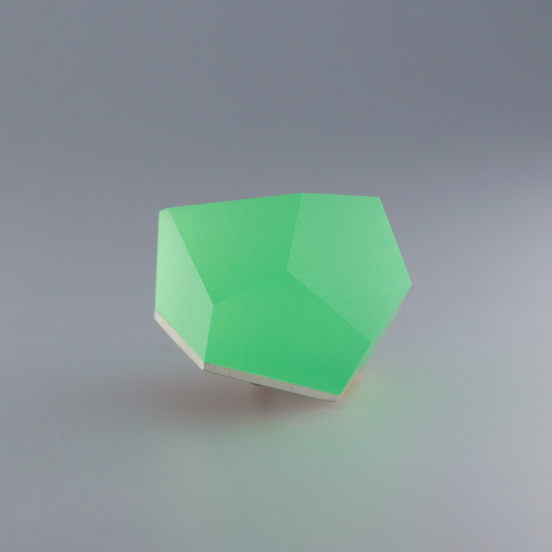 A geometric green gemstone with a faceted, irregular polyhedral shape sits against a plain grey background. The stone's surface is smooth and slightly translucent, with a softly glowing appearance, making it an exquisite Fruit Bijoux Ring Top VU Crystals Flash Green in sterling silver or 24ct gold-plated settings.