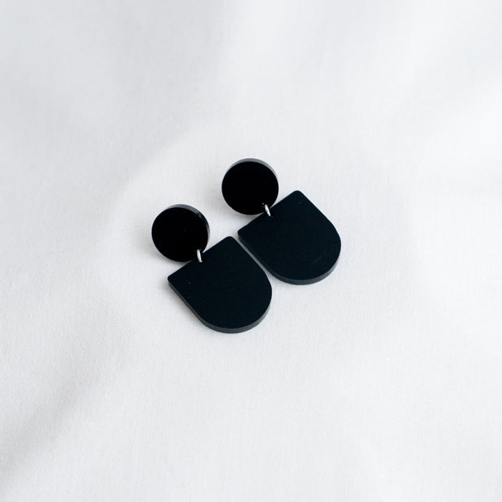 A pair of Silver 14 Black Earrings by Studio Nok Nok lies on a light fabric background. Each earring features a round top connected to a larger, flat, and rounded rectangular bottom piece, creating a clean and modern geometric design. These handmade earrings are crafted from recycled acrylic for an eco-friendly touch.