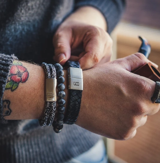 Close-up of a person's wrist adorned with several bracelets: a multicolored braided leather bracelet with a stainless steel clasp, Steel and Barnett's Preston - Matt Black beaded bracelet, and another genuine leather bracelet. The person is wearing a grey sweater and has a colorful flower tattoo on the wrist.